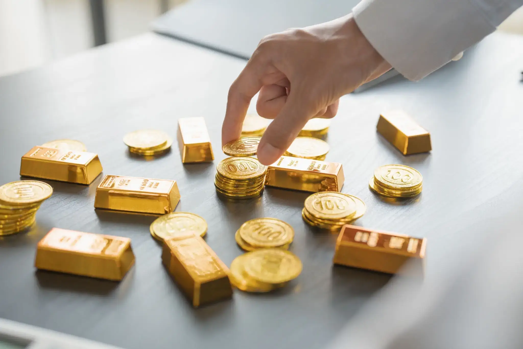 Hand stacking gold coins and bars on a table.